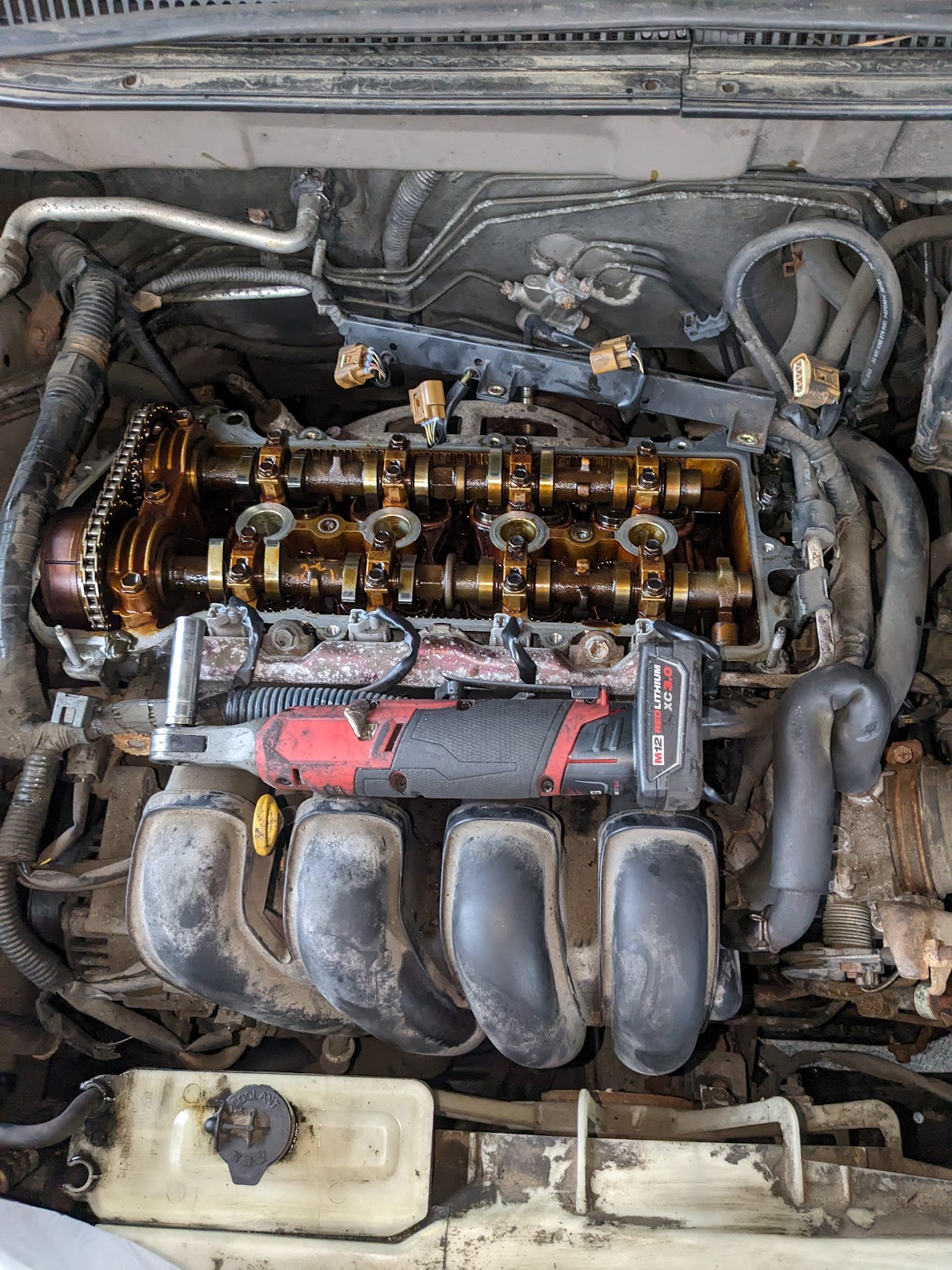Robert's Mobile Services — four-cylinder engine with valve cover removed exposing camshafts, a Milwaukee M18 impact driver staged mid-service